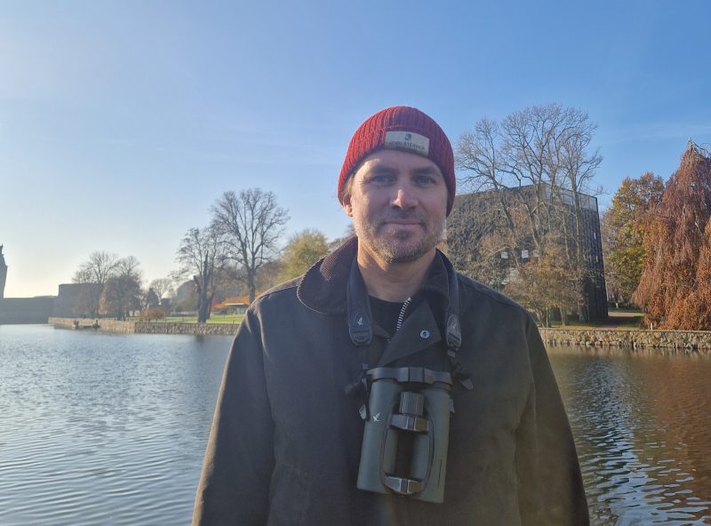 En man tittar in i kameran och ler. Runt halsen har han en kikare. Himlen är blå och i bakgrunden skymtas Kalmar konstmuseum.