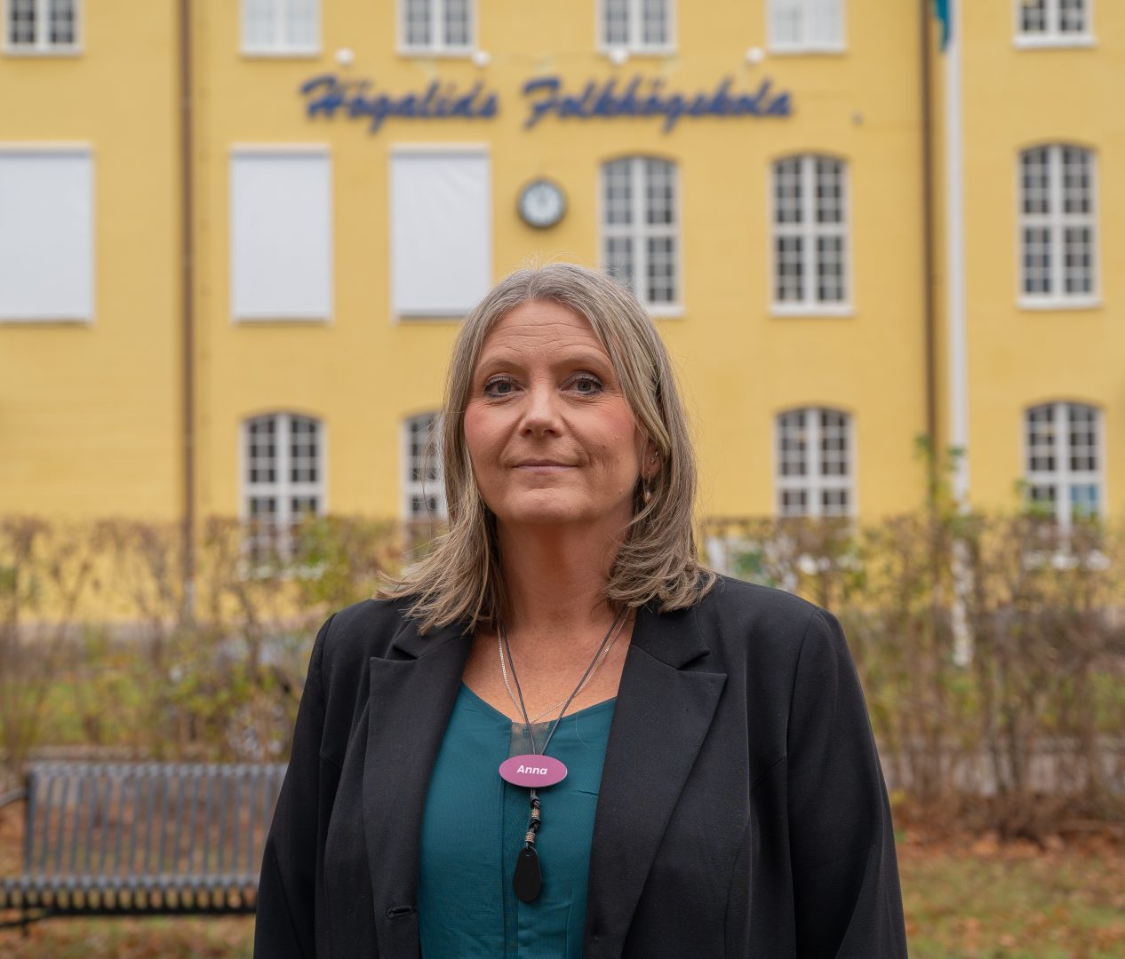 En kvinna står utomhus framför en gul byggnad med skylten "Högalids Folkhögskola". Hon har axellångt blont hår och bär en mörk kavaj. På bröstet har hon en namnskylt med texten "Anna".
