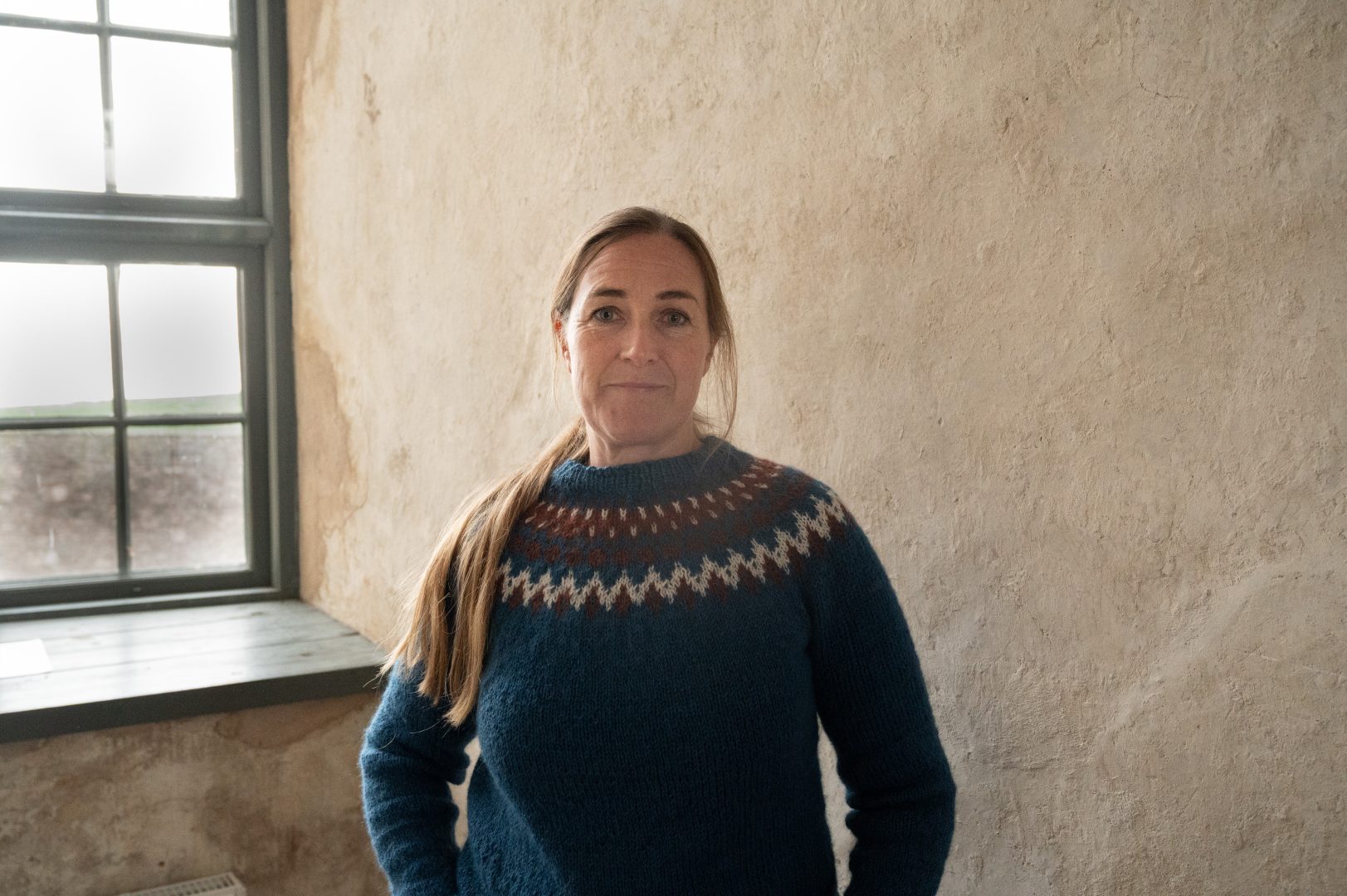 En kvinna i blå tröja bredvid ett fönster. Fönstret och väggarna är synligt gamla.