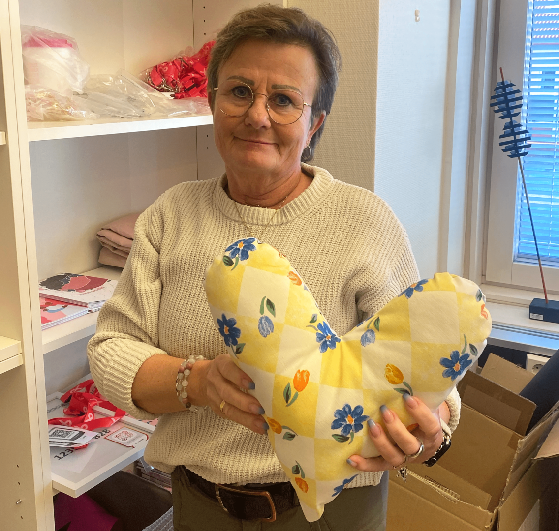 Kvinna framför en hylla med produkter från Bröstcancerföreningen. Kvinnan håller en hjärtformad kudde.