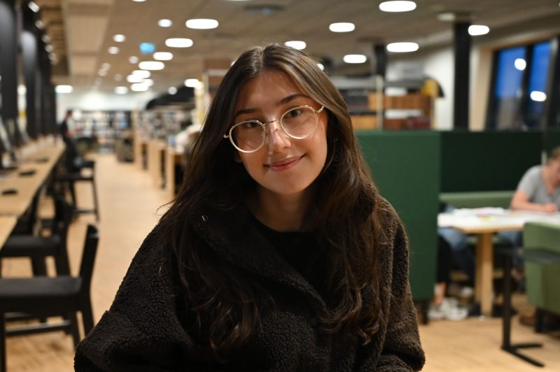 Linnéa Widegren sitter med sin bruna jacka och silvriga glasögon på en stol i universitetsbiblioteket Kalmar. Bakom henne sitter en grupp människor i gröna soffor och studerar. Bakgrunden är suddig med Linnéa i fokus.