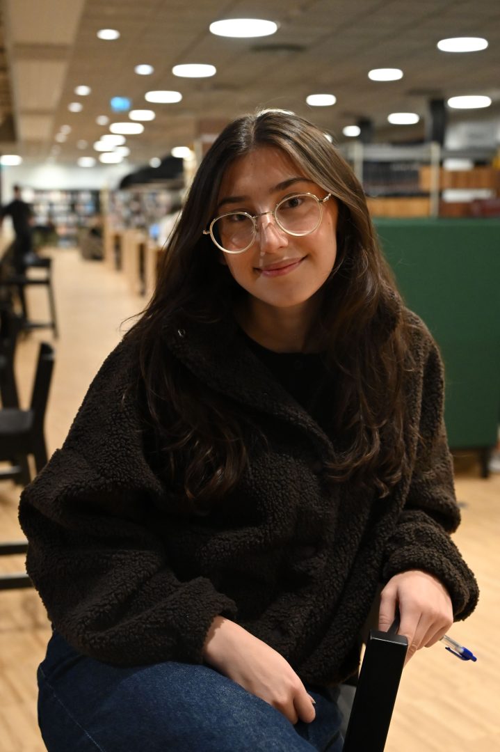 En helbild på Linnéa Widegren i en svart stol mitt i Universitetsbiblioteket Kalmar. Bakgrunden är suddig och framhäver Linnéa i mitten av bilden. Hon har en brun teddyjacka, svarta jeans och håller i en blå bläckpenna. I bakgrunden syns gröna soffor och fler svarta stolar.