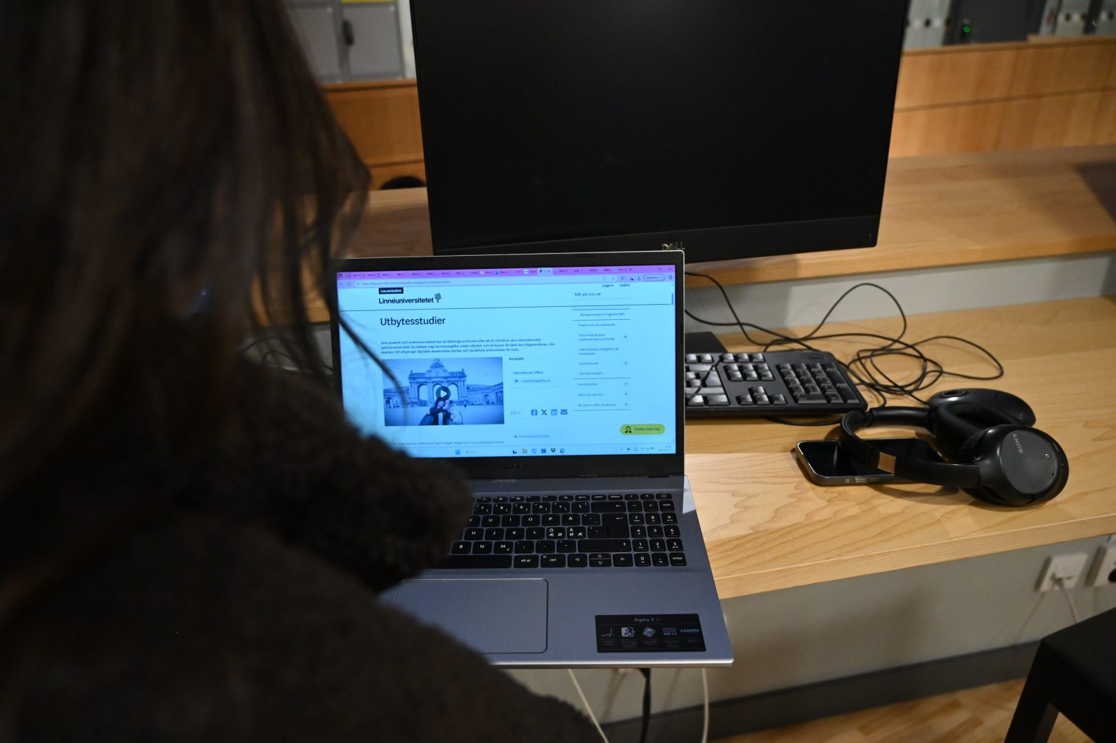 En bild tagen över Linnéa Widegrens axel där en dator syns. På datorn står det om utbytesstudier hos Linnéuniversitetet. Bredvid datorn ligger en svart telefon och svarta hörlurar.