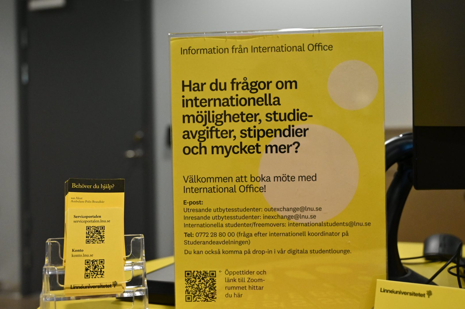En gul plastskylt med vita prickar med svart text om internationella frågor och möjligheter. Informationen är från International Office. Brevid den gula skylten finns kort från Linnéuniversitetet med svarta Qr koder.