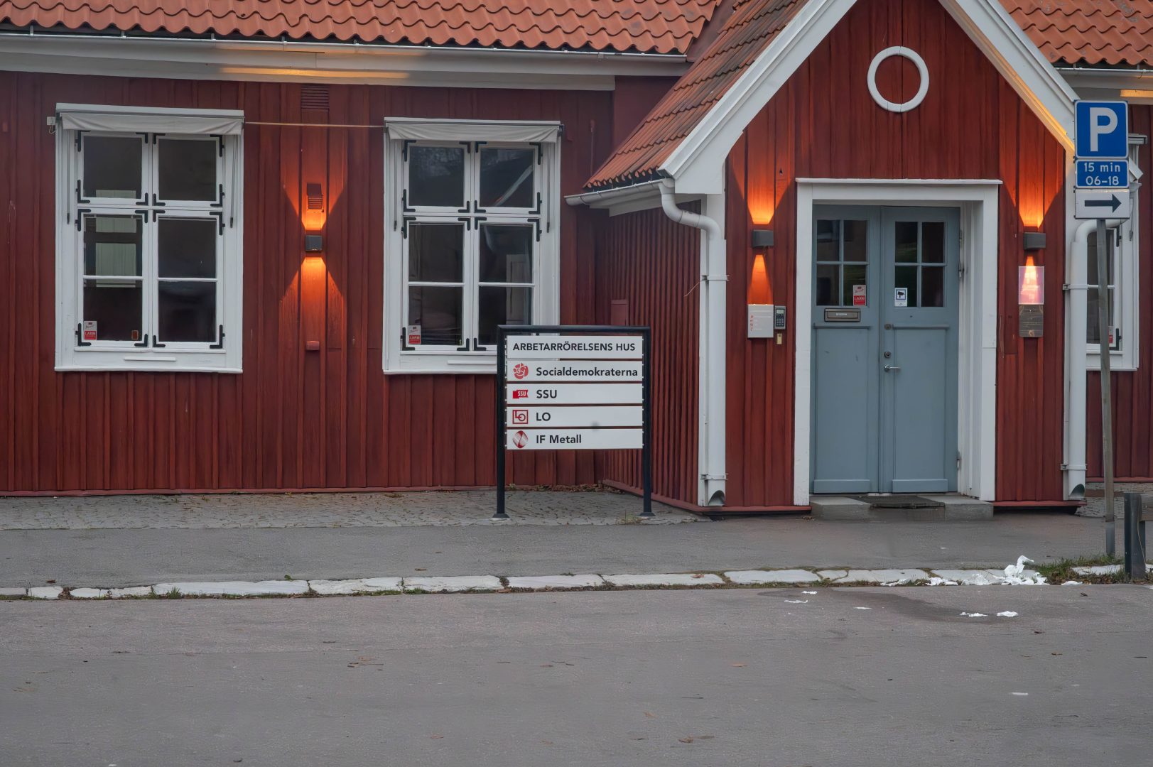En röd trähusbyggnad med vita fönsterkarmar och en grå dörr. Framför byggnaden står en skylt med texten ’Arbetarrörelsens hus’ och logotyperna för Socialdemokraterna, SSU, LO och IF Metall. Till höger om dörren finns en parkeringsskylt som anger 15 min parkering mellan 06–18.