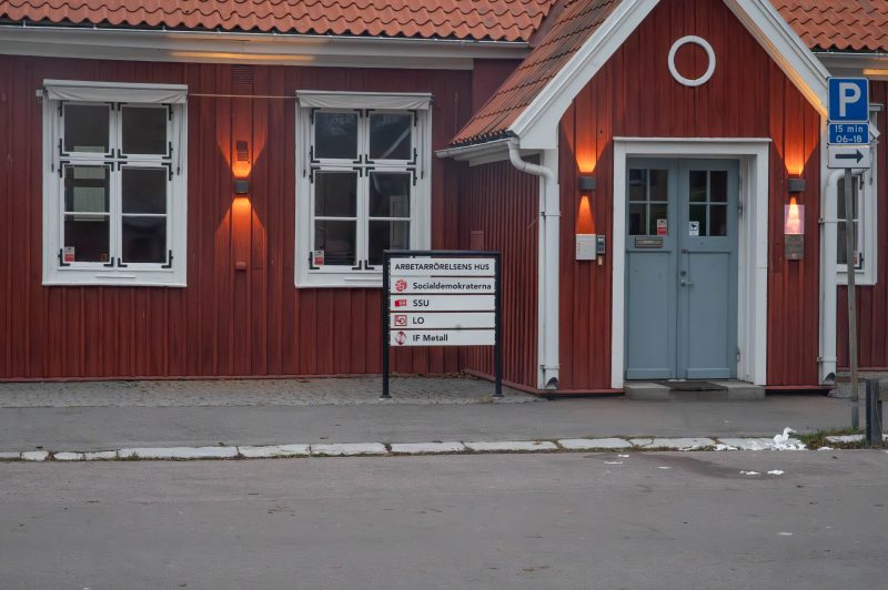 En röd trähusbyggnad med vita fönsterkarmar och en grå dörr. Framför byggnaden står en skylt med texten ’Arbetarrörelsens hus’ och logotyperna för Socialdemokraterna, SSU, LO och IF Metall. Till höger om dörren finns en parkeringsskylt som anger 15 min parkering mellan 06–18.