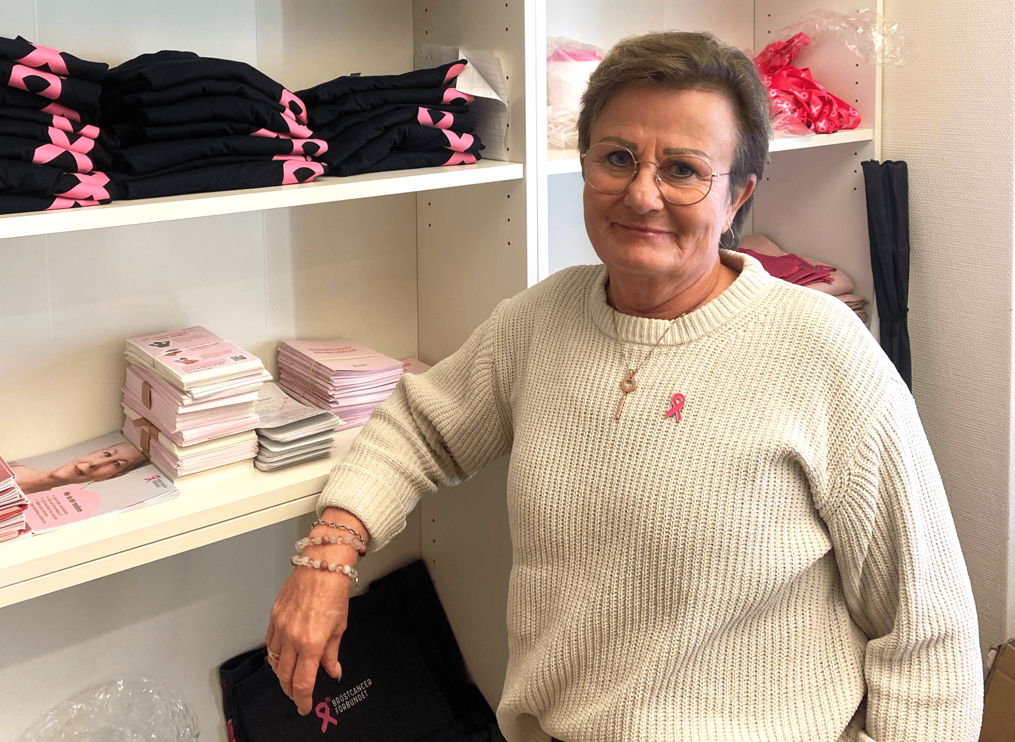 En kvinna framför en hylla med produkter från Bröstcancerföreningen. På tröjan har hon ett rosa band.