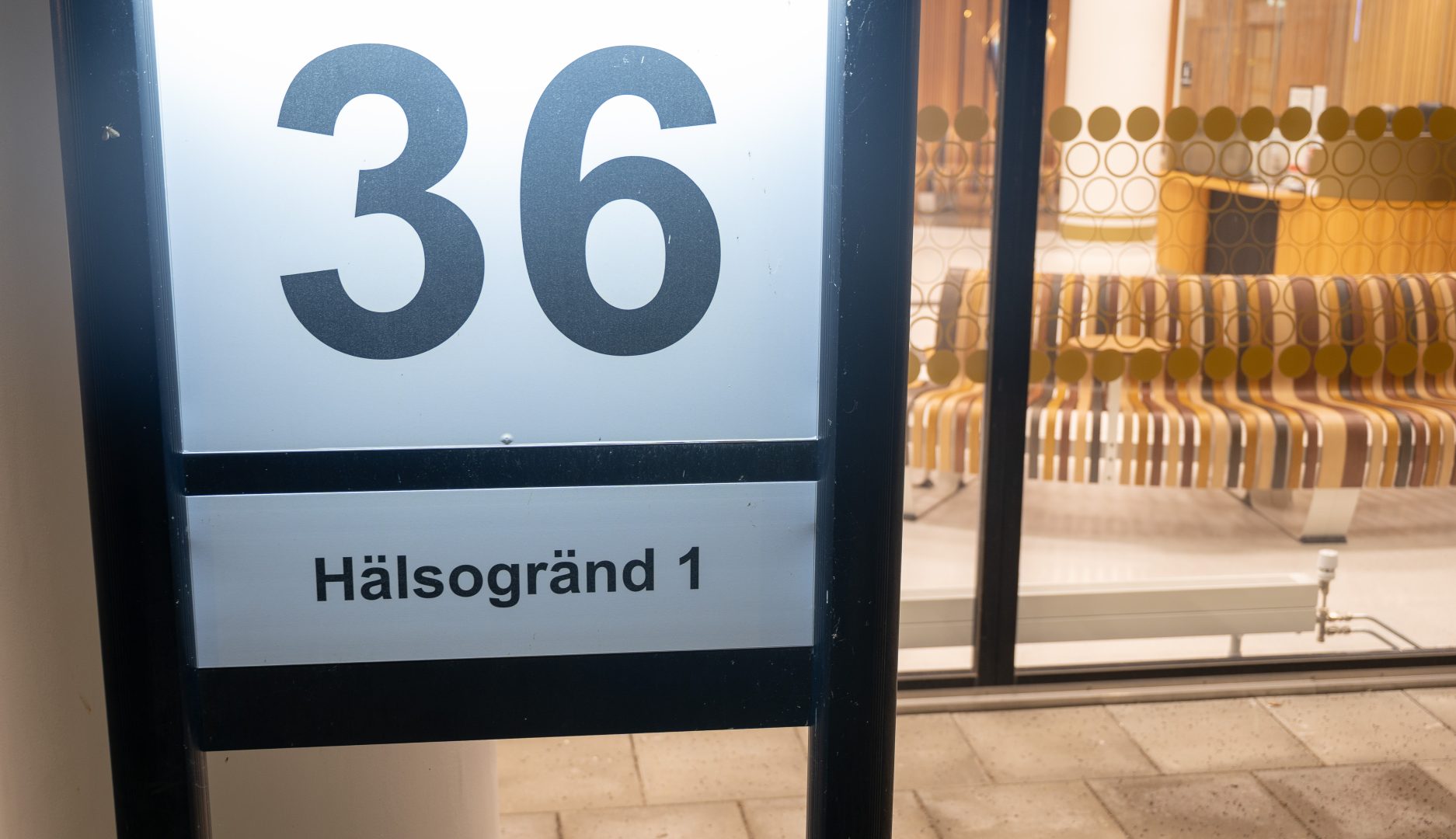 Närbild av en upplyst skylt med siffran 36 och texten "Hälsogränd 1". I bakgrunden syns bland annat randiga bänkar och ett lågt element.