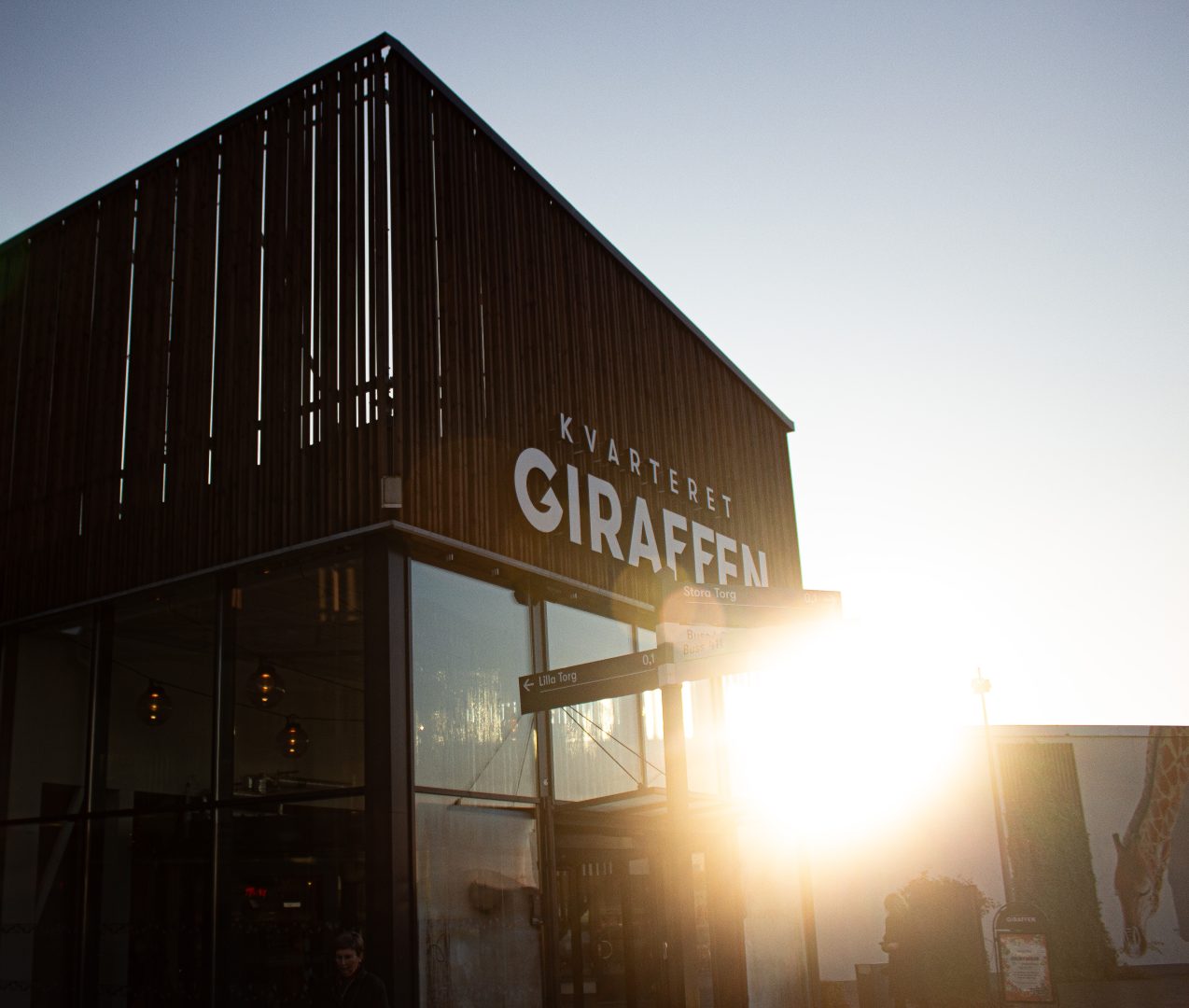 Ett modernt byggnadshörn i trä och glas med starkt solsken som lyser in från sidan. På fasaden står det ”Kvarteret Giraffen”. Framför entrén finns en skylt med riktningar och i bakgrunden syns en målning av en giraff på en vägg. Foto: Juwelia Jansson