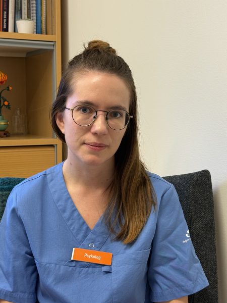 En person med långt hår och glasögon sitter inomhus, klädd i blå vårduniform. Hon bär en namnbricka med texten "Psykolog". I bakgrunden syns en bokhylla med böcker och några dekorationer.