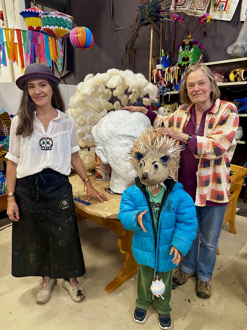 Organisatörerna står nära en stor mask och ler mot kameran. Nära Bodil Göransson står ett barn med en djur mask