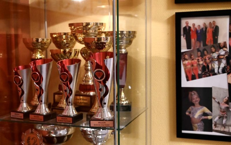 På bilden sys ett flertal pokaler i ett glasskåp. På väggen bredvid hänger bilder med minnen från danstävlingar.