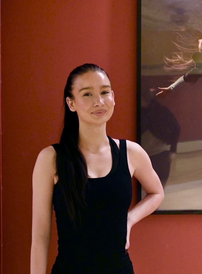 På bildens syns en ung kvinna klädd i ett svart träningslinne och håret uppsatt i en hög hästsvans. Hon står mot en röd vägg. På ena sidan av bilden syns även en del av en stor tavla med en dansande kvinna som motiv. 