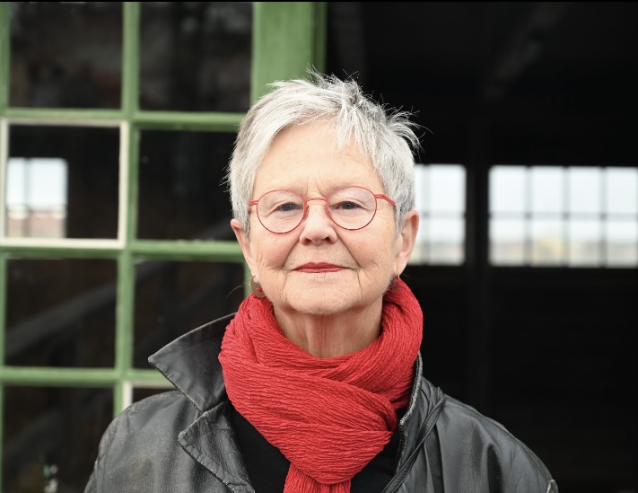 En kvinna med kort blont hår står framför en halft öppen dörr. Hon ler. Hon bär röda glasögon, rött läppstift och en röd halsduk.