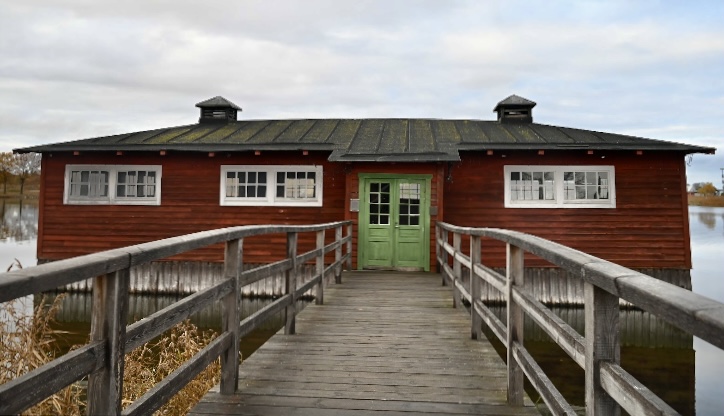 Bilden visar hela huset. Ett mellanstort rött hus. I förgrunden syns en mörk trä bro med räcken. Vid sidorna syns vatten och vass. 