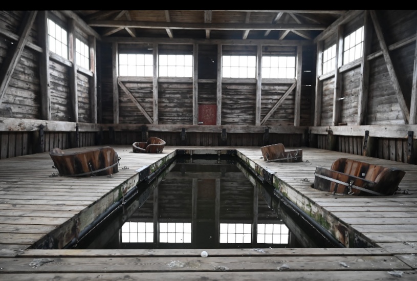 Bilden är tagen innefrån tvättguset och färgerna går i gråa toner. I mitten är en rektangulär basäng omgiven av ett flertal baljor i trä. Både golv och väggar är i slitna plankor. Ljus släpps in längst med sidorna va fönster