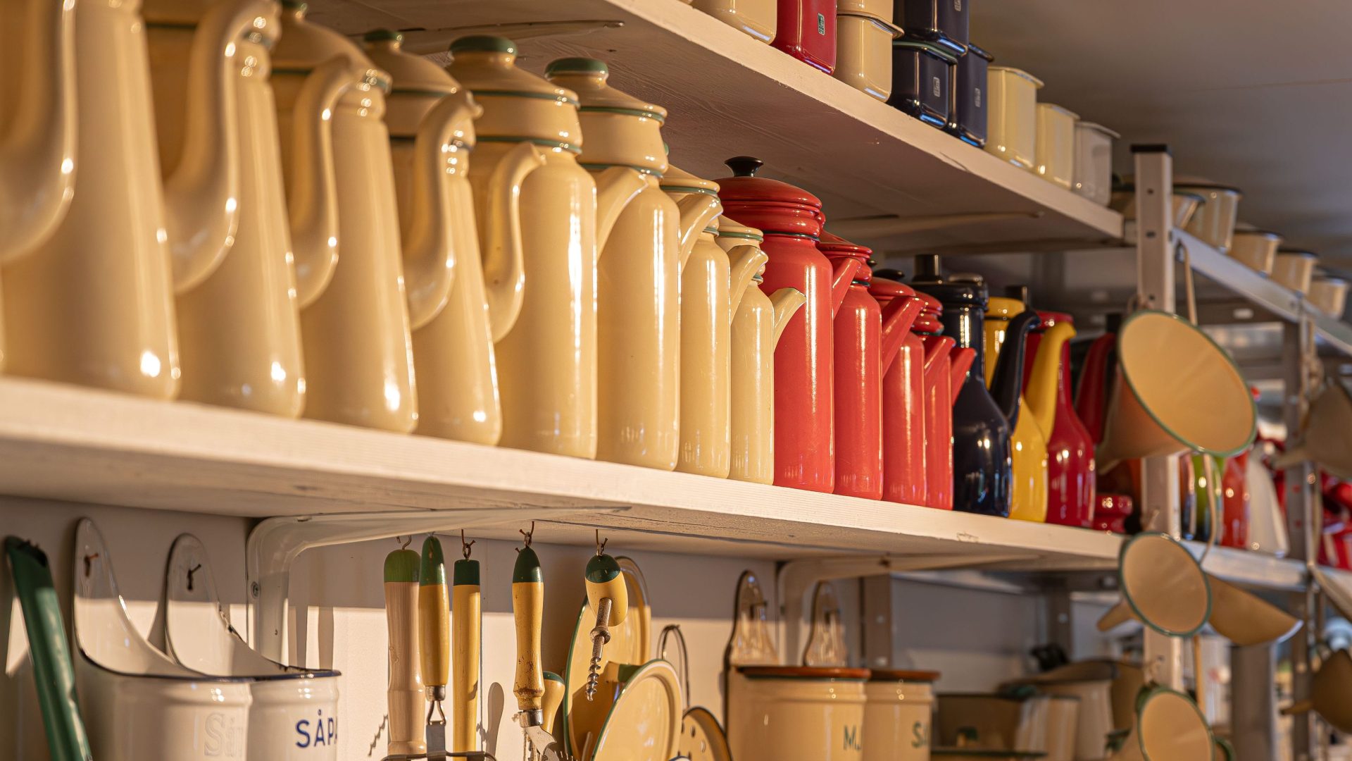 En vägg med flera hyllor fyllda med gamla emaljerade kökskärl i olika färger och former. På den främre hyllan står rader av beige kaffekannor, följt av röda, mörkblå och gula kannor längre bort. Under hyllorna hänger köksredskap med träskaft och gröna detaljer, samt durkslag och behållare märkta med text som ”SÅPA”. Ljuset från sidan ger ett varmt sken över de blanka ytorna, vilket skapar en nostalgisk känsla av ett gammaldags kök.. 