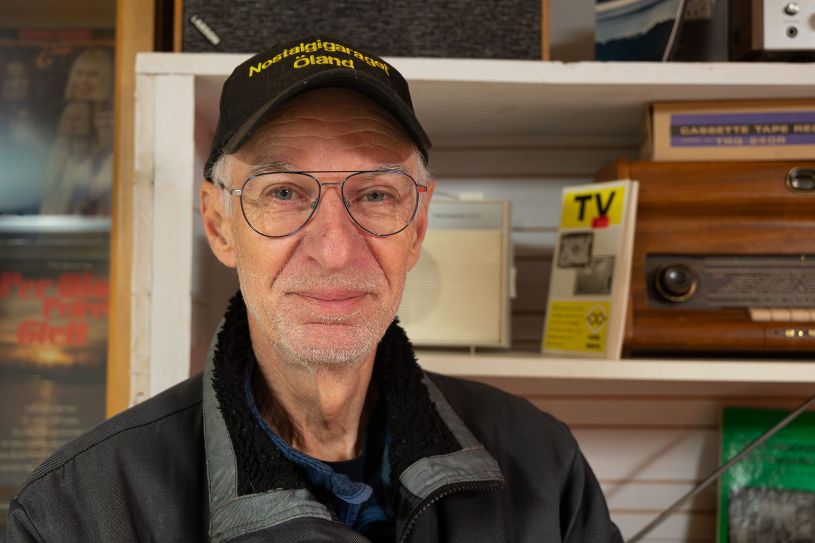 Porträttbild av Lars Karmlid som står frmför en bakrund av äldre radioaparater och broschyrer. Lars är iklädd en svart jacka, glasögon och en keps med texten "Nostalgigaraget Öland"
