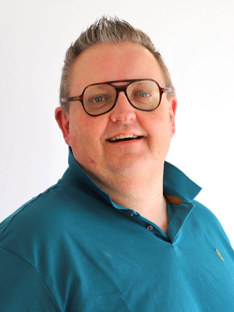 En man med kort, gråsprängt hår och glasögon ler mot kameran. Han bär en turkos pikétröja med uppvikt krage och står mot en vit bakgrund.