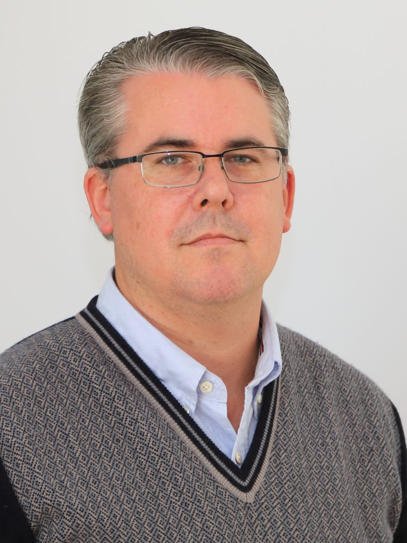 n man med kort, gråsprängt hår och glasögon ser rakt in i kameran med neutral min. Han bär en ljusblå skjorta under en grå och mörkblå stickad väst med mönster. Bakgrunden är vit.