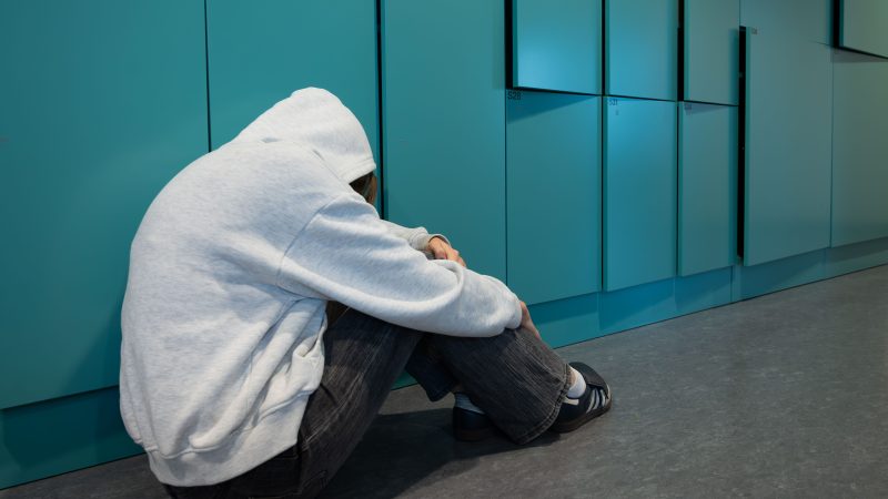 Tjej med huva sitter på golvet framför skåp i en skolkorridor. Hon ser ledsen ut.