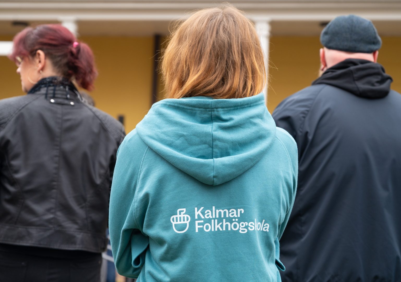 En person med ljusbrunt hår står med ryggen mot kameran och bär en turkos tröja med texten "Kalmar Folkhögskola" på ryggen. Två andra personer står bredvid.
