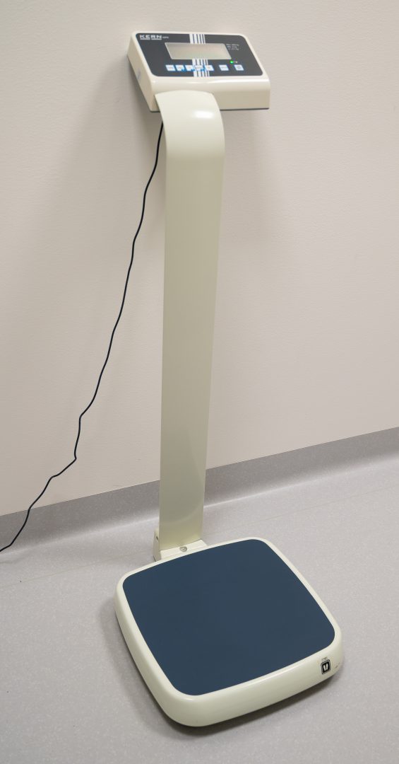 En digital personvåg med en upphöjd display monterad på en böjd stång. Vågen står på ett ljust golv intill en vägg, och en strömkabel hänger ned från displayen.