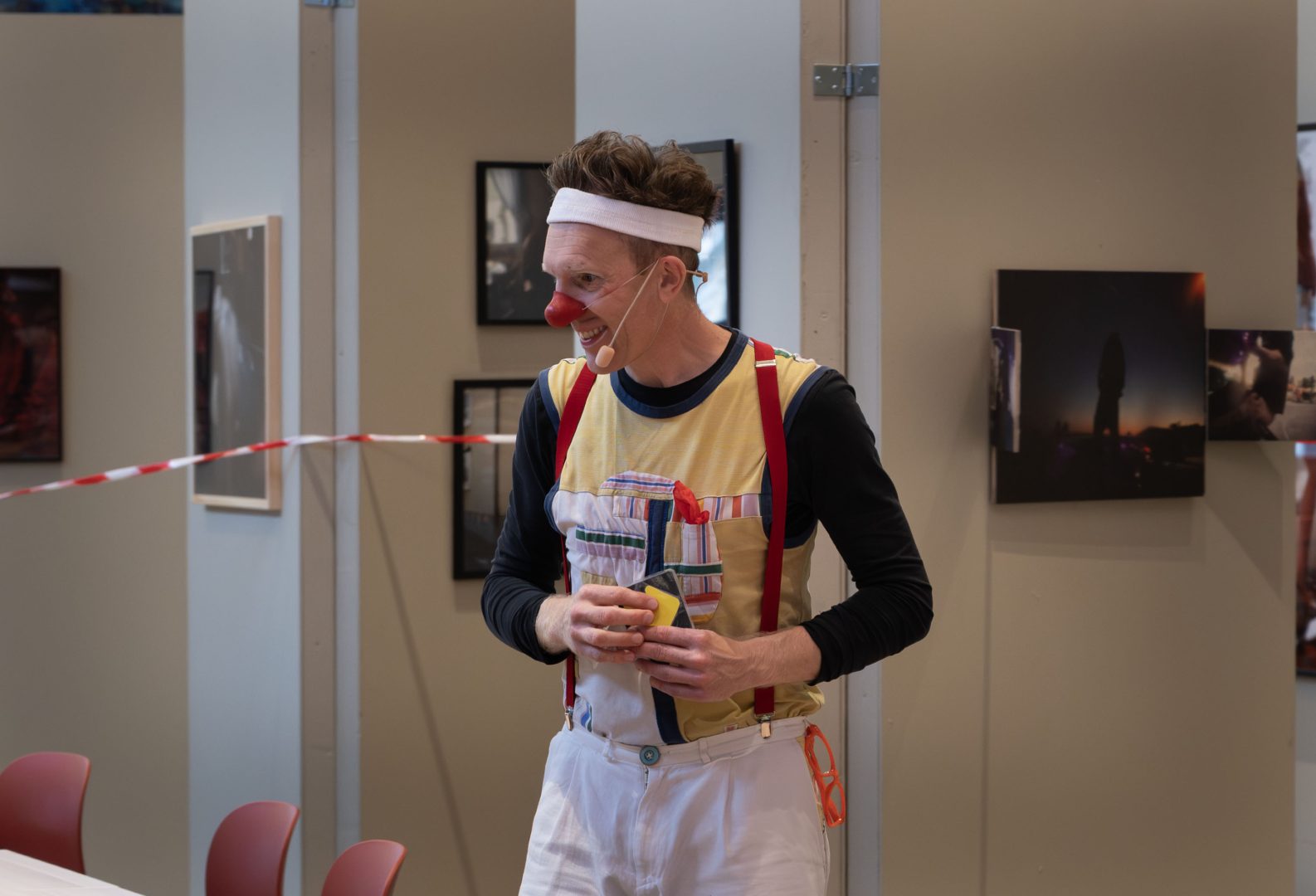 En clown står inomhus med ett leende. Han bär en röd clownnäsa, vit pannband, gula hängslen, och färgglada kläder med röda detaljer. Han håller något i händerna och verkar mitt i en uppträdande eller lekfull stund. I bakgrunden syns tavlor på väggarna och några röda stolar.