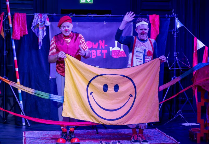 Två clowner står på en scen under starkt färgat blått och lila ljus. Den ena clownen bär röd basker, röd väst, röd fluga och rödvitrandig tröja, medan den andra har stora röda hängslen, vitt svettband och färgglada kläder. De håller tillsammans upp en stor gul flagga med ett stort svart smiley-ansikte på. Den ena clownen vinkar glatt mot publiken, och färgglada tygband och flaggor pryder scenen runt dem.