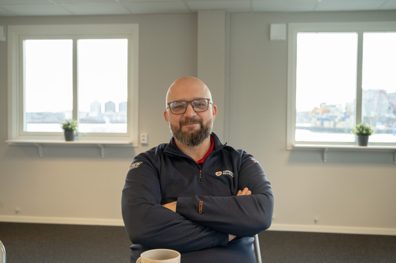 bild på man med glasögon sitter ner med armarna i kors. Två fönster på vardera sida. 