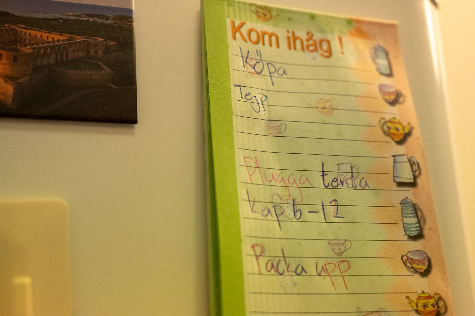 En grön, orange Kom ihåg! lista sitter på ett vitt kylskåp. På lappen står det köp tejp, plugga tenta kap 6-12 och packa upp. På listan finns även olika kaffemuggar i flera färger. Bredvid listan till vänster sitter en magnet halvt synlig av Borgholms slott.
