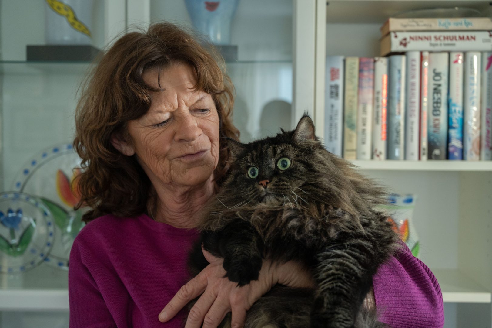 Carina Olsson håller sin bruna, fluffiga katt i famnen. Hon tittar på den och hennes lila tröja lyser upp bilden. Kattens gröna ögon tittar åt vänster. Bakom dem finns en bokhylla med glas och böcker.