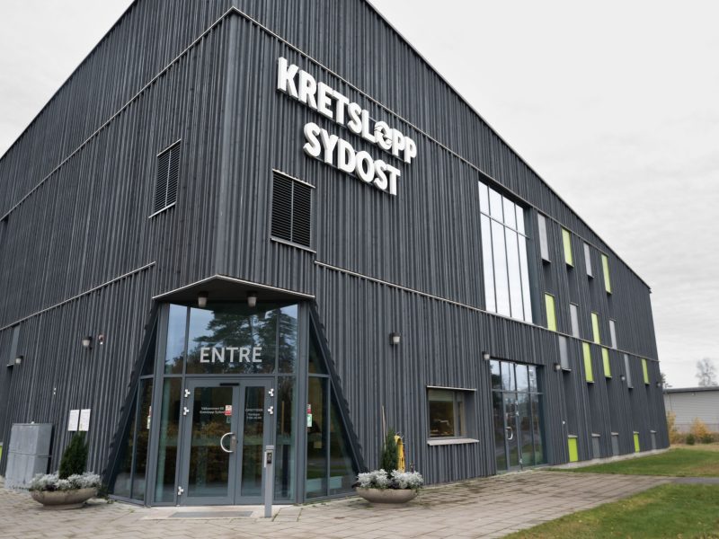 På bilden är det en grå modern byggnad med gröna detaljer och Kretslopp Sydost är skrivet på fasaden. I hörnet av byggnaden är det en entre till byggnaden där det står ordet entre över. Utanför entren är det två blomkrukor med vita blommor i.