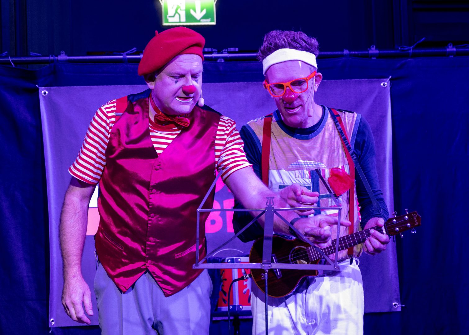 Två clowner står på en scen under ett framträdande. Den ena clownen bär en röd basker, röd väst, röd fluga och en röd-vitrandig tröja. Den andra clownen har stora orange glasögon, ett vitt svettband, färgglada kläder och håller en ukulele. De står tätt intill varandra och tittar på något på ett notställ. Bakgrunden är upplyst i blått och lila ljus, och en grön nödutgångsskylt syns ovanför dem.