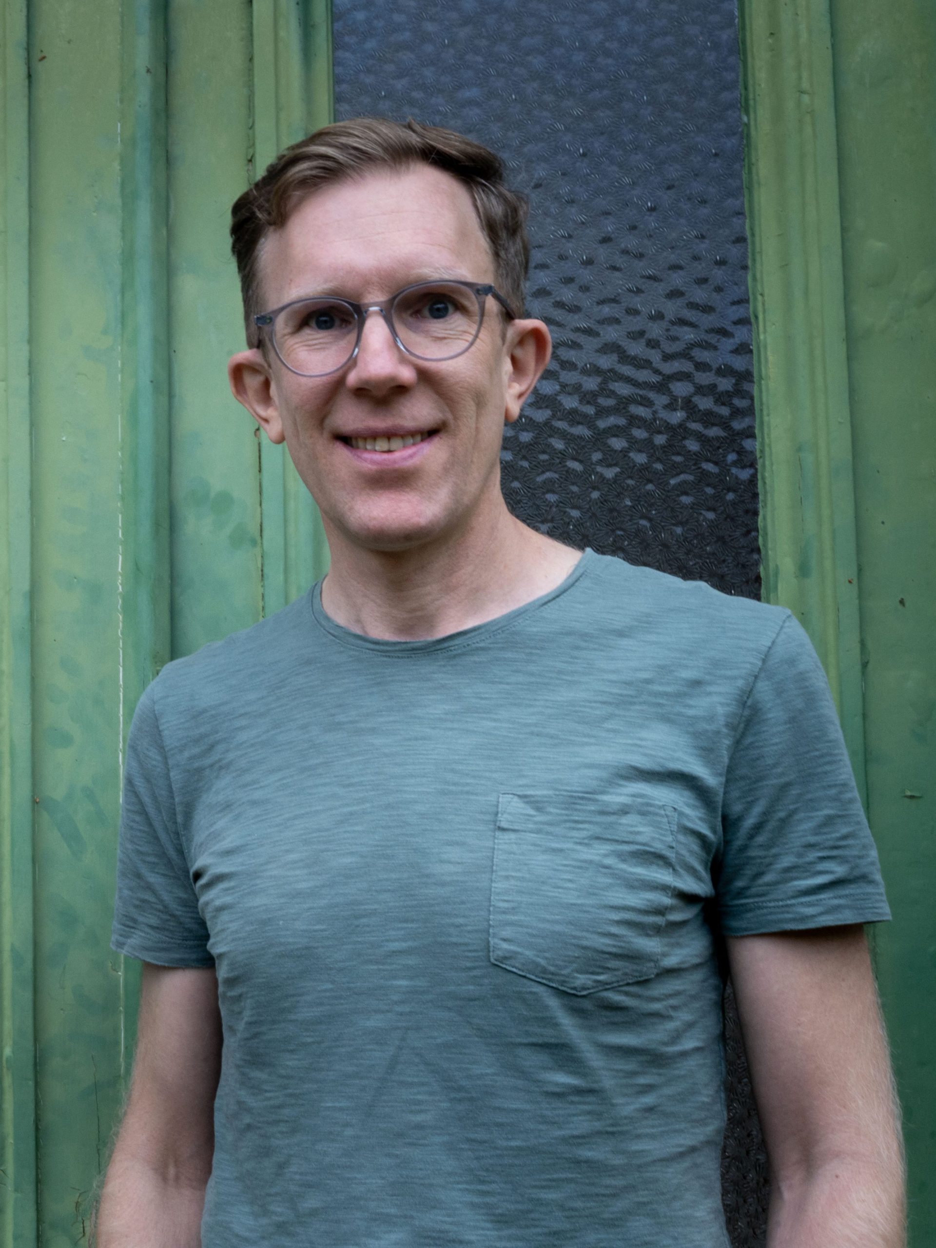 En person med glasögon och kort hår står utanför och ler framför en grön vägg med en glasruta.