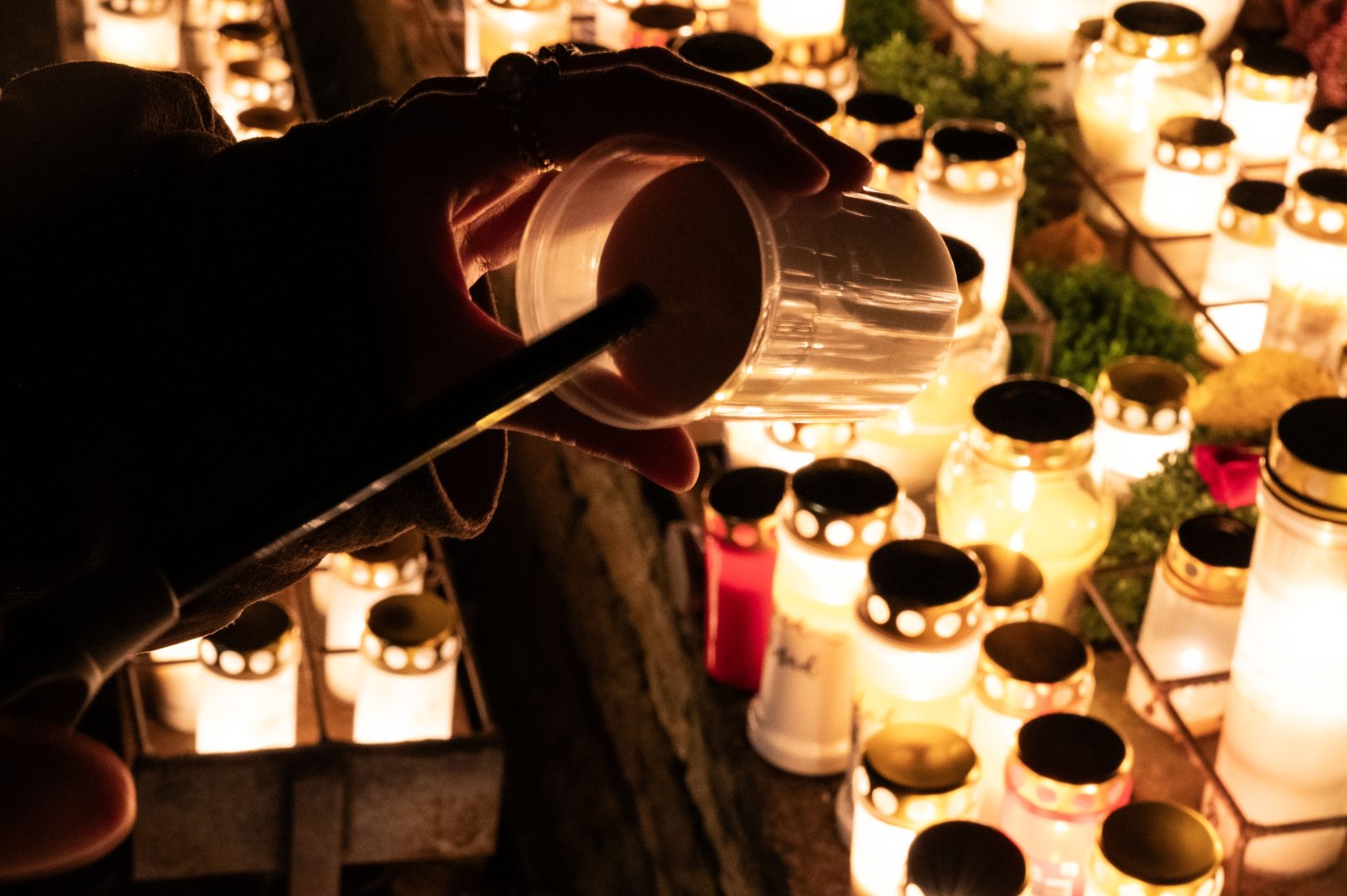 En hand i skugga som med en tändare håller på att tända ett gravljus. I bakgrunden ser man flertalet fler tända gravljus.