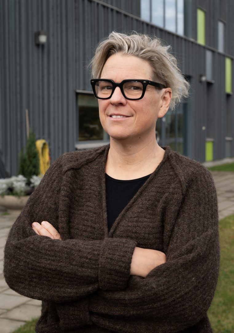 En person står utomhus framför en modern byggnad med grå fasad och gröna fönsterdetaljer. Personen har kort, ljust hår, bär svarta glasögon, en mörkbrun stickad tröja och en svart tröja under. Hen har armarna i kors och ler lätt mot kameran.