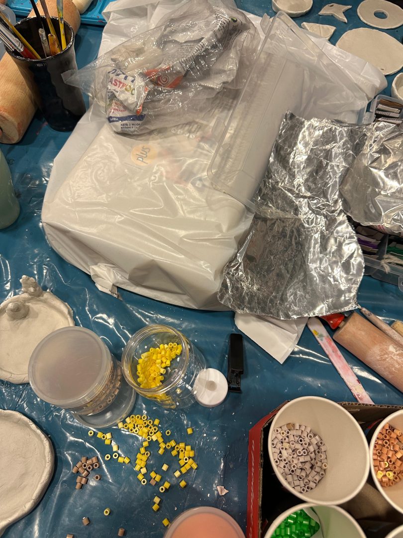 Inplastad lera ligger på ett inplastat bord. Runt omkring står burkar och pappersmuggar med pärlor i.